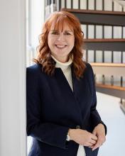 Woman with red hair wearing a navy blazer and white turtleneck stands smiling by a window, with shelves of binders in the background.