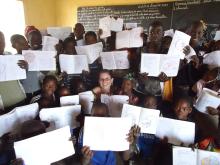 A group of children and an adult sit and stand in a classroom, holding up notebooks with drawings. A chalkboard with writing is visible in the background.