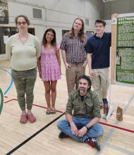 Five people pose by a science poster presentation in a gymnasium; four stand while one sits on the floor in front of the group.