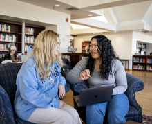 Two women sit on armchairs in a library, having a conversation. One holds a laptop on her lap, while bookshelves and another person are visible in the background.