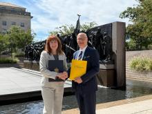 Two people in business attire shake hands outdoors in front of a large bronze monument, each holding official-looking folders.