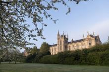 Large stone building with pointed towers sits behind a green lawn and blooming trees under a clear blue sky.