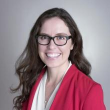 A woman with long brown hair wearing glasses, a white top, and a red blazer smiles at the camera.