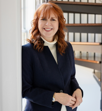 A smiling woman with shoulder-length auburn hair wearing a navy blue blazer over a cream-colored turtleneck.