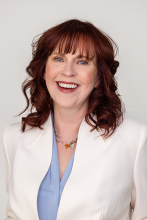 A smiling woman with shoulder-length reddish-brown hair wearing a light blue top and a white blazer.