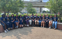 A group of fourteen young people wearing matching dark blue t-shirts with a gold circular graphic sit in a row on a low brick wall.