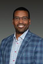 A smiling man wearing glasses and a plaid blazer over a light-colored button-up shirt.