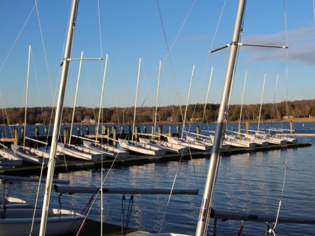 Angled photo of sailboats stationary on the floating docks, without sails. The sky is bright blue and the water is calm. 