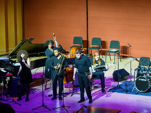 Students perform on stage on piano, brass, bass, drums and signing