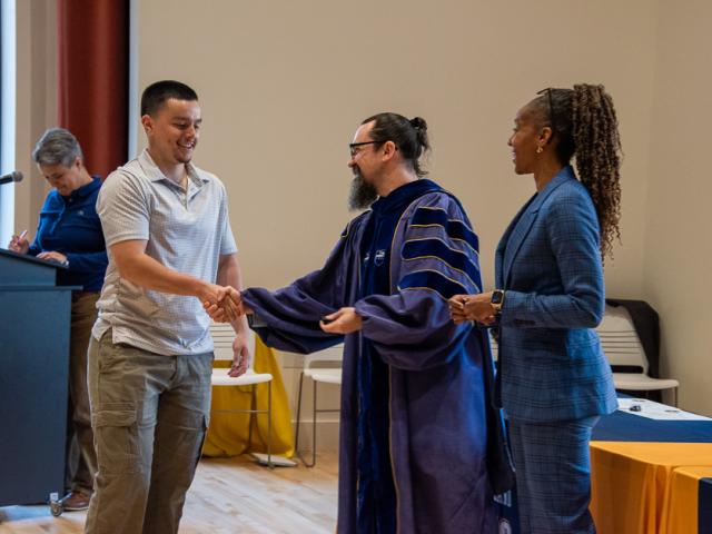 Man shakes hands with another man alongside a bystander in a large room