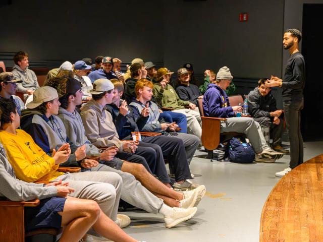 Speaker in a darkened theater with seated audience applauding