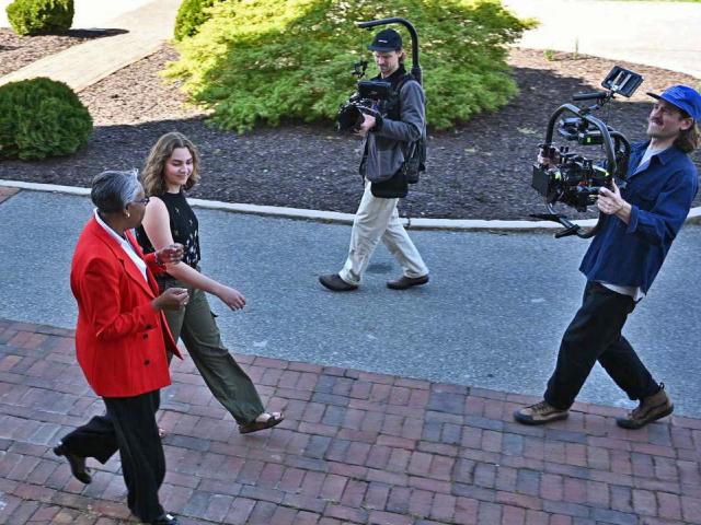 Two people walk in front of two video cameramen