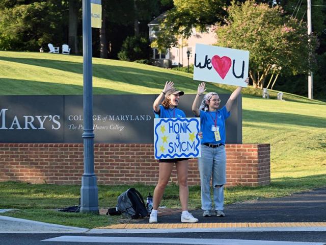 Students holding welcome signs wave to cars at campus entrance