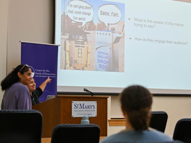 two people at a podium point to a meme on a big screen as the audience looks on. 