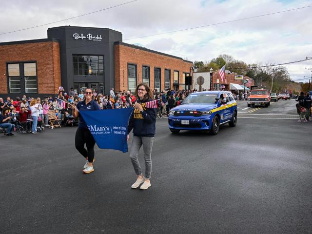 Two women carrying SMCM banner march in parade in front of SMCM Public Safety cruiser