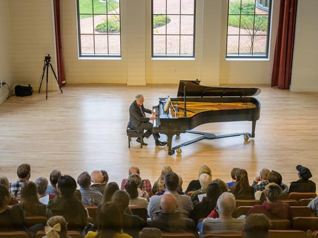 Man seated at piano before crowd in recital hall - video camera filming to the left