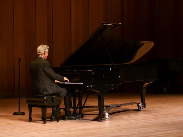 An adult with gray hair, dressed in a dark suit, is playing a grand piano on a wooden stage. A microphone stand is positioned to the left of the person, and the background features wooden paneling.