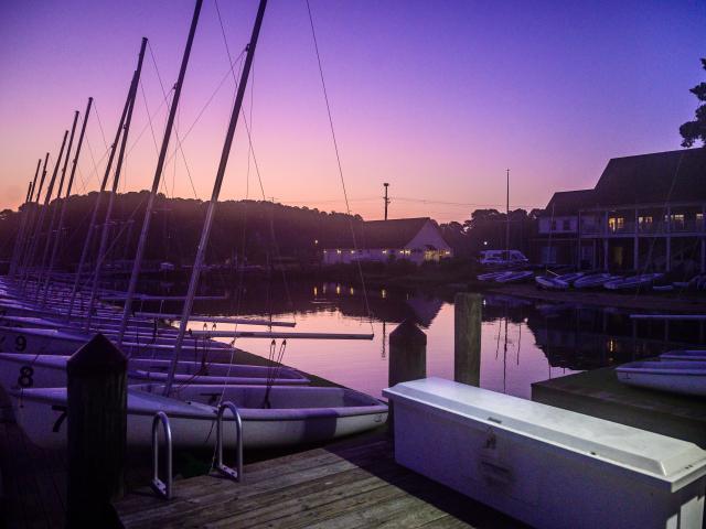 A serene marina at twilight with sailboats docked on the calm water. The sky transitions from orange to purple, reflecting on the water. Buildings and trees are silhouetted in the background. A white bench is on the wooden dock.