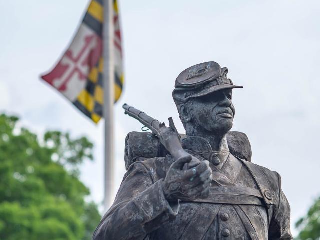 Statue of a soldier in uniform holding a gun against his chest, looking upwards. Behind the statue, a flagpole with the Maryland state flag is visible. The sky is overcast, and trees are in the background.