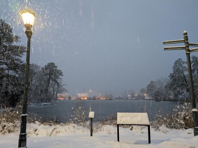 Snow falls gently on a serene lakeside. A glowing streetlamp lights the scene, with trees and a wooden sign covered in snow. In the background, distant lights reflect on the water, creating a peaceful winter atmosphere.
