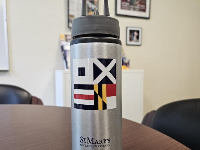 Silver water bottle with a dark lid on a round table. The bottle features colorful flag patterns and the text "St. Mary's College of Maryland." Blurred background includes framed posters and photos on a wall.