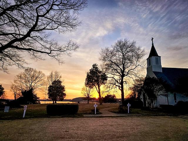 A serene scene of a church with a steeple silhouetted against a colorful sunset sky. Bare trees frame the foreground, and a body of water and hills are visible in the distance.