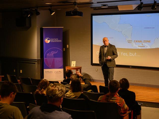 A person stands on a small stage giving a presentation to an audience. A projector screen shows a map including the Panama Canal Zone. A banner is visible on the left side of the stage. People are seated and facing the speaker.