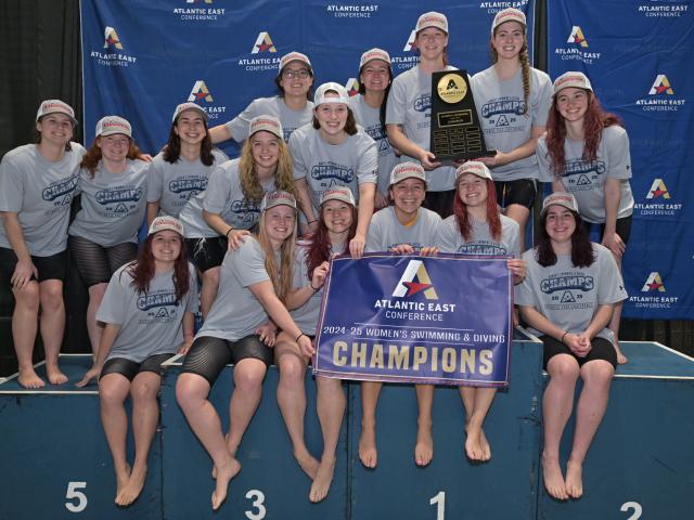 A women's swim team poses together wearing matching shirts and caps, holding a trophy and a banner that reads "Atlantic East Conference 2024-25 Women's Swimming & Diving Champions" against a branded backdrop.