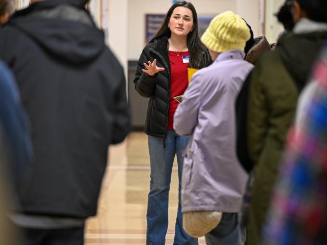A woman stands in a hallway, gesturing with one hand as she speaks to a group of people. She has long dark hair, wears a red top and blue jeans, and has a name tag. The group is listening attentively, some wearing winter clothing.
