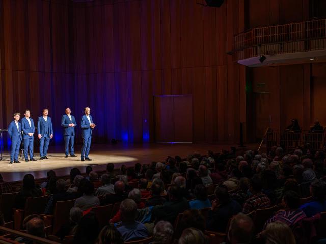 A group of six people in blue suits sing on a lit stage in a large concert hall. An audience fills the seats, watching attentively. The hall features wooden walls and a balcony.