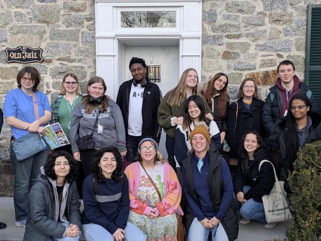A group of people posing in front of a stone building with a white door and a sign that reads "Old Jail." Some are standing, and others are crouching. The group appears diverse in terms of gender and ethnicity. Vegetation is visible in the foreground.