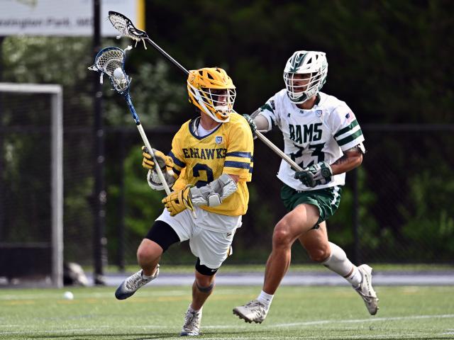 Two male lacrosse players are on a field during a game. The player in yellow and blue, wearing number 2, runs with the ball under pressure from the player in white and green, wearing number 13. A net and trees are visible in the background.