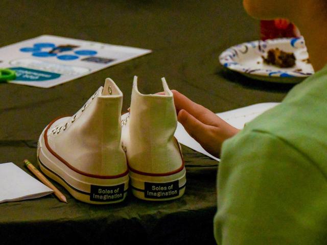 A person seated at a table with a pair of white high-top sneakers labeled "Soles of Imagination," scissors, a paper plate with cake, and papers. The focus is on the sneakers and the person’s hand resting beside them.