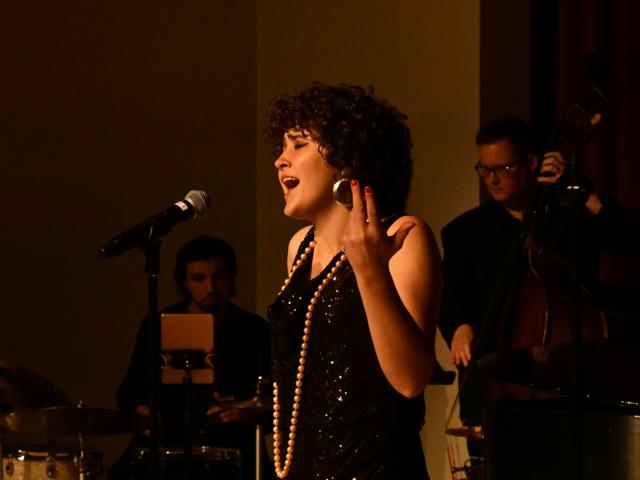 A woman with curly hair sings passionately into a microphone, wearing a black sequined dress and pearl necklace. A band plays in the background with a drummer and a double bassist. The scene is warmly lit, creating a cozy atmosphere.