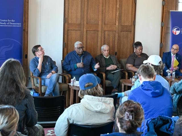 A panel of five people seated in chairs, engaged in a discussion. An audience listens attentively in a room with banners displaying "Center for the Study of Democracy." The panelists are diverse in appearance and age.