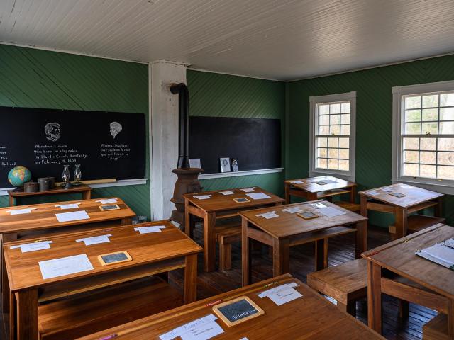 A vintage classroom with wooden desks, a blackboard displaying chalk drawings and text, old-fashioned stove, and educational posters. The room is lit by natural light from the large windows, showcasing a historical school setting.