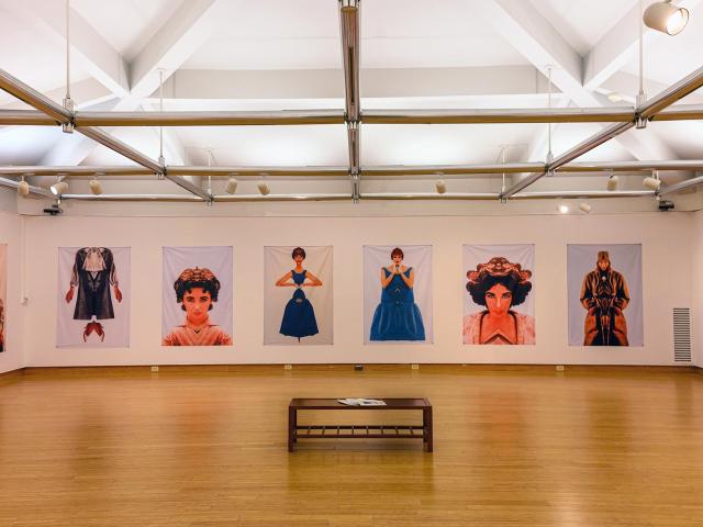 A spacious art gallery features large portraits of individuals in different poses. The images are evenly spaced on white walls. A wooden bench with a magazine sits in the center of the hardwood floor under bright lighting.