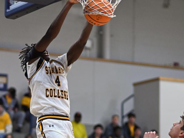 A basketball player wearing a white and gold "St. Mary's College" uniform, number 4, performs a slam dunk. Another player in a red jersey is visible below. Spectators in yellow shirts sit in the blurred background.