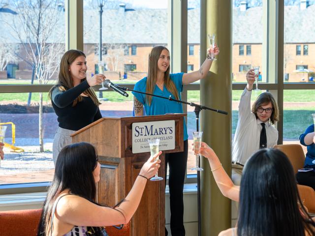 A group of people raising glasses in a toast at St. Mary's College of Maryland. Two women are at a podium, smiling, and holding microphones. Others are seated around, participating in the toast. Large windows and a brick building are in the background.