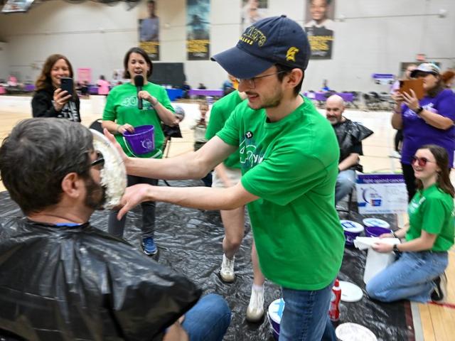 A young man in a green shirt playfully smashes a whipped cream pie into another man's face at an indoor event. The recipient wears glasses and a black plastic cover. Nearby, people watch and laugh, wearing casual clothing.