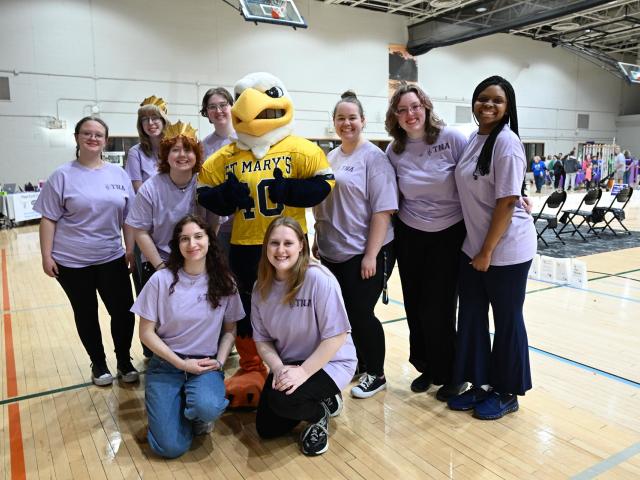 A group of nine people stand smiling indoors on a wooden sports court. They are wearing matching light purple shirts and are grouped around a mascot dressed as a bird in a yellow jersey with "ST. MARY'S" on it.