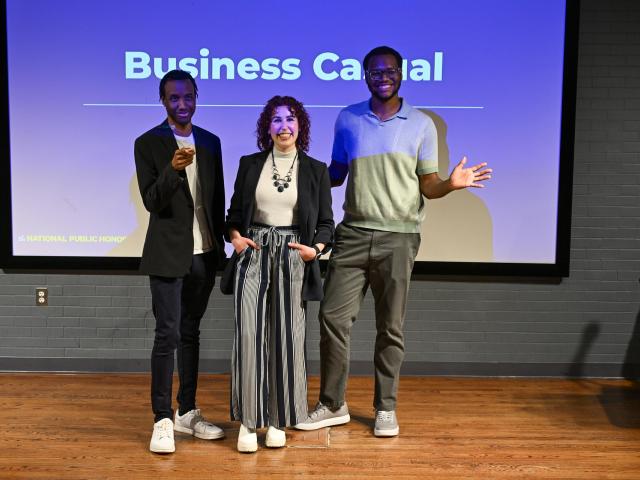 Three people stand smiling in front of a presentation screen displaying "Business Casual." They are dressed in casual professional attire with sneakers. The background features a dark brick wall and wooden floor.