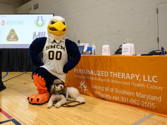 A mascot wearing a "SMCM" jersey poses with a dog in front of a table with an orange cloth from Personalized Therapy, LLC, in a gym setting.