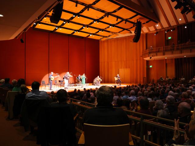 A musical performance is taking place on a well-lit stage in a large auditorium. Musicians play various instruments as a full audience watches. The walls and ceiling are wooden, enhancing the room's acoustics.