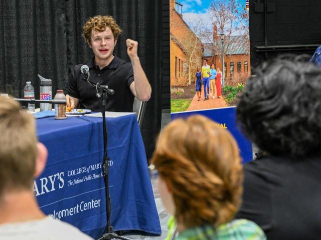 A young person with curly hair speaks emphatically at a microphone, seated at a table with a blue tablecloth. An audience listens attentively. A large poster with a group of people walking outdoors is visible behind them.