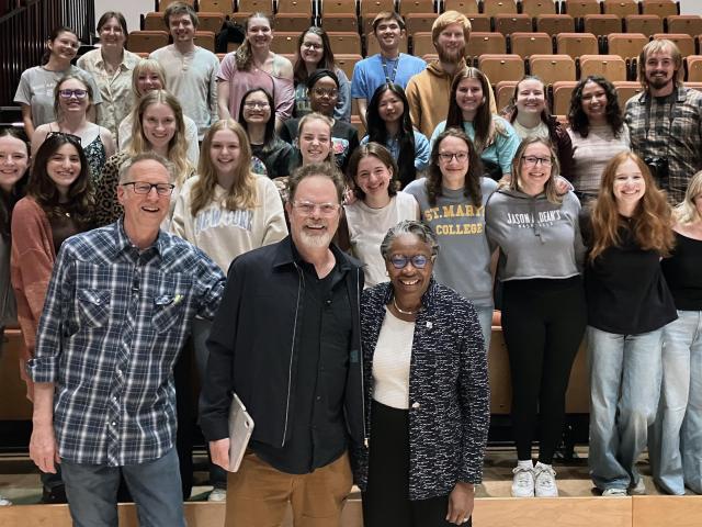 A group of people, including three in front, pose for a photo in a lecture hall with stadium seating.