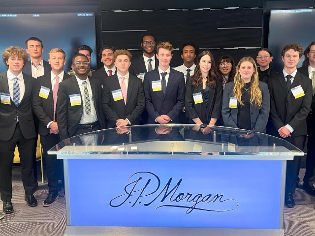 A large group of people dressed in business attire stand behind an illuminated J.P. Morgan desk. Two screens are visible in the background. The room has a corporate, professional setting.