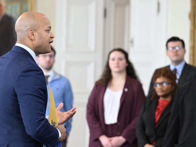 A man in a blue suit speaks to a group of people indoors. The group includes individuals in formal attire, standing attentively. A portrait hangs on the wall in the background, and there's natural lighting in the room.