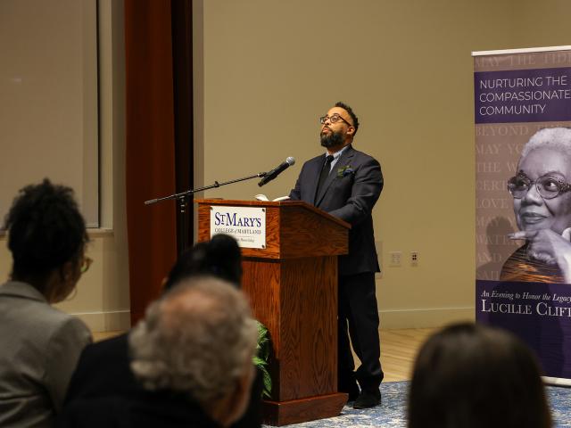 A man stands at a podium giving a speech in a room with a banner featuring Lucille Clifton that reads "Nurturing the Compassionate Community." Audience members are seated in the foreground.