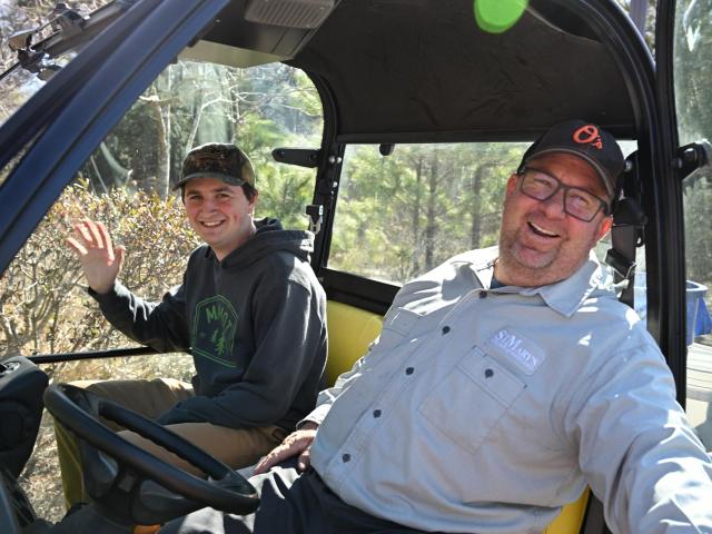 Two men sit in a vehicle, facing the camera. The man on the left waves and wears a cap and hoodie. The man on the right, also in a cap, smiles broadly. They appear relaxed and outdoors, surrounded by trees and bushes.